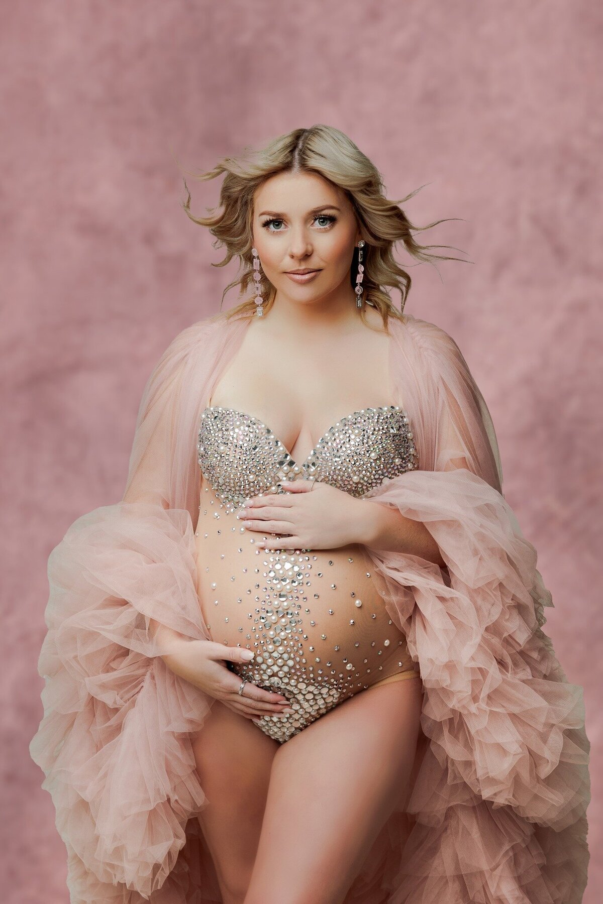pregnant mother wearing rhinestone sheer one piece with a blush pink tulle wrap holding her baby belly bump