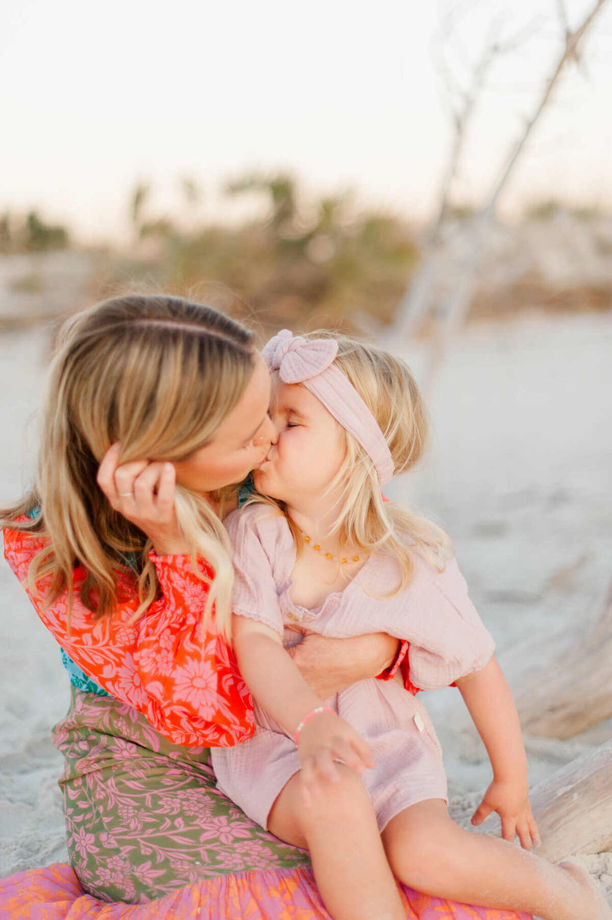 Young daughter kissing her mom while sitting in the sand at sunset