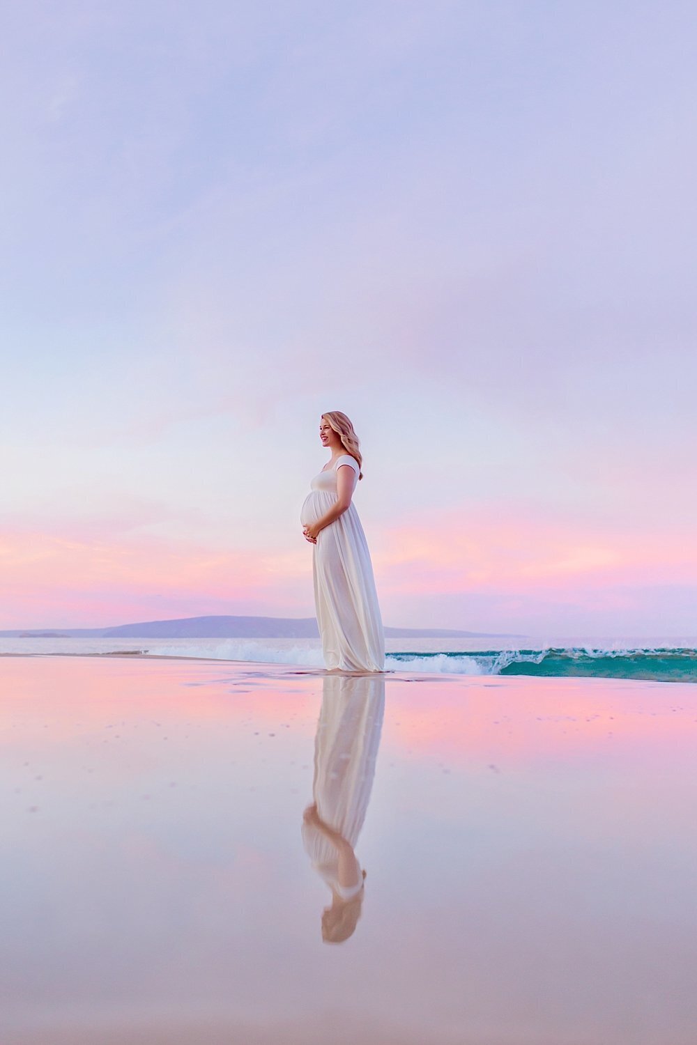 Blonde woman wearing a white silk maternity dress holds her tummy at sunrise, one of the best times of day for Maui maternity portraits