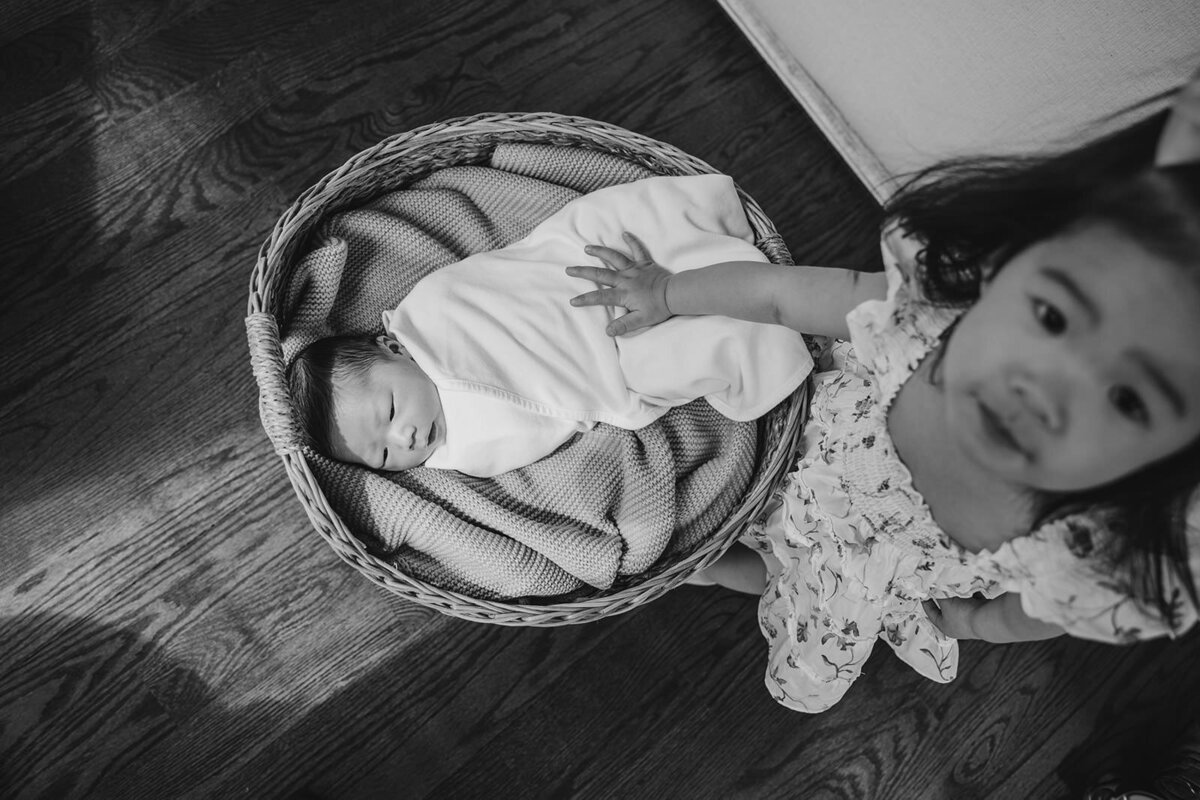 A small chile reaching out to touch their newborn sibling in a basket 