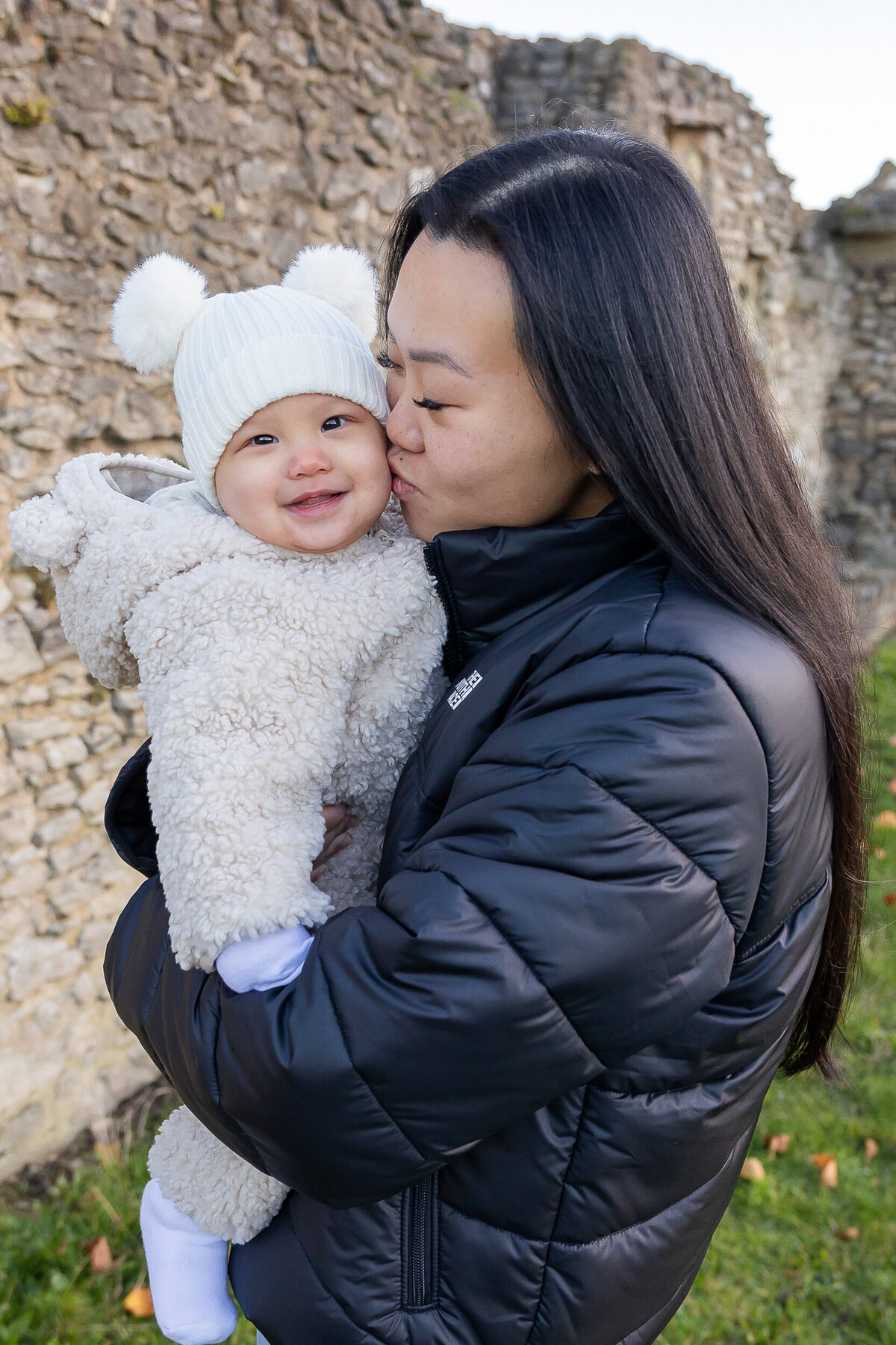 A mother lovingly kisses her baby, who is smiling and dressed in a fluffy white outfit and hat, against a backdrop of ancient stone ruins.