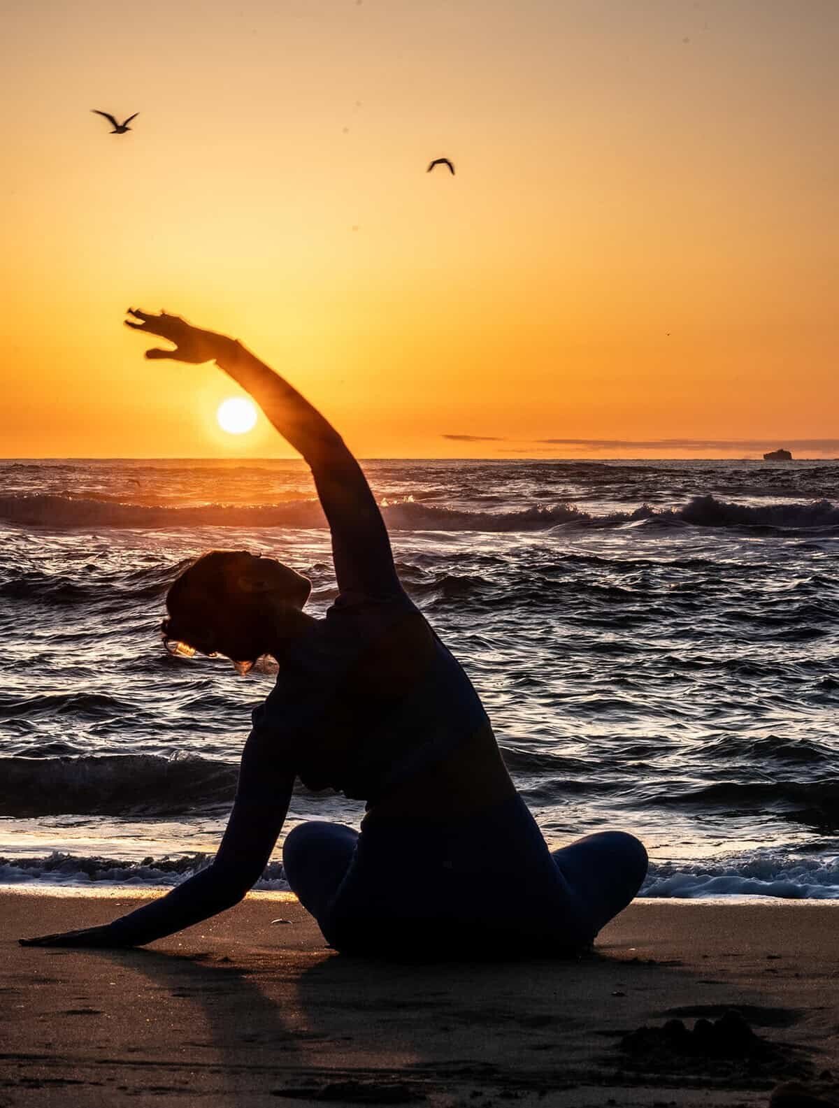 Woman in a yoga pose sitting and stretching while watching the sun set over the ocean