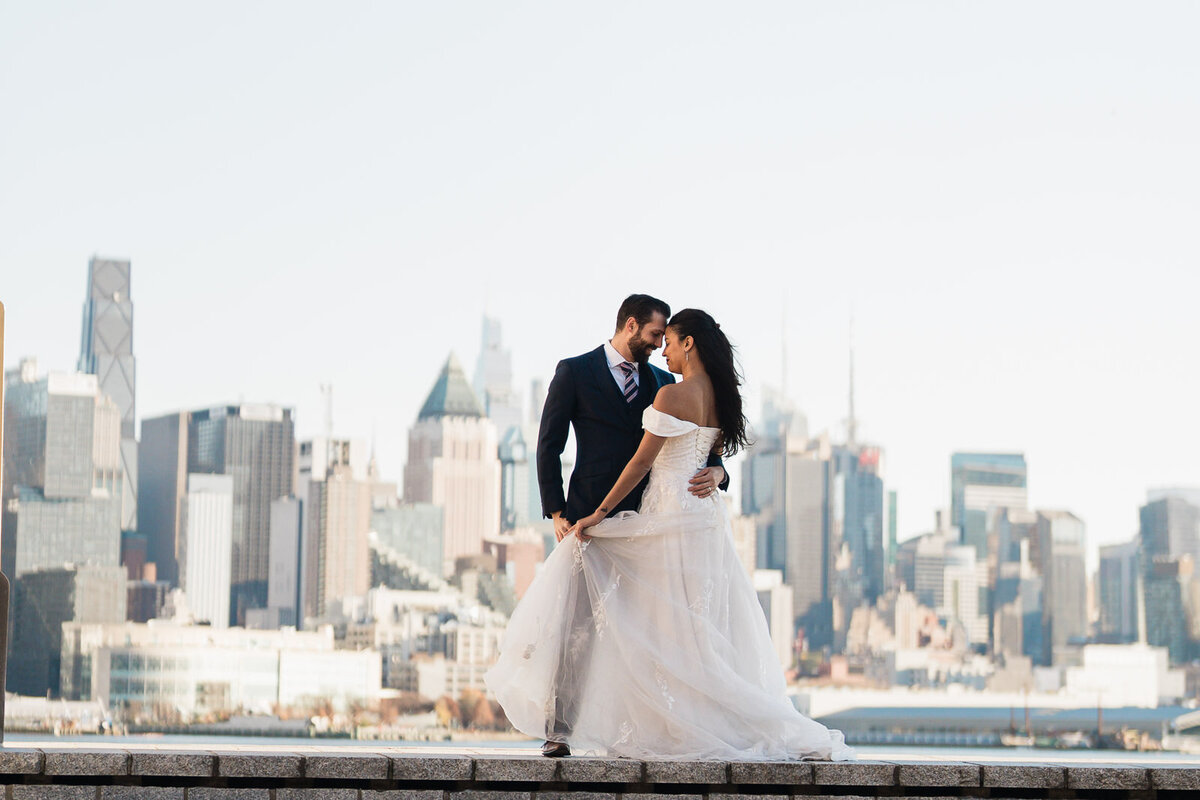 Bride and groom standing on the Hoboken waterfront with the New York City skyline behind them, sharing a romantic moment during their wedding portrait
