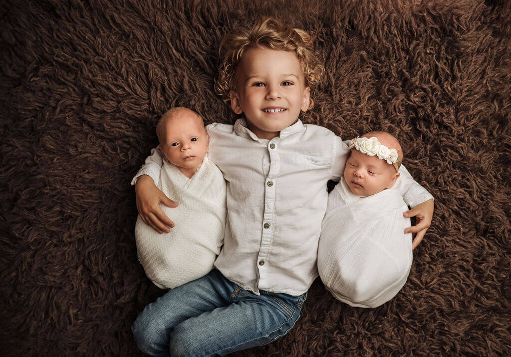 Toddler big brother holding his brother and sister newborn twins wrapped up laying on a fur rug smiling at the camera.