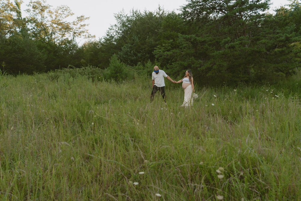 A cinematic photo of a couple walking through a grassy field. The man is leading his pregnant wife while she carries a bouquet of flowers in Johnson City, TN. 