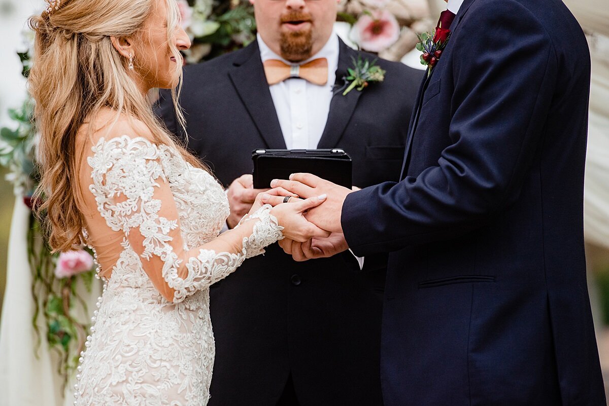 Standing in front of a floral arbor, the blonde bride, wearing a long sleeved lace wedding dress with a plunging back holds hands with the groom, wearing a dark blue suit and burgundy boutonniere as the minister in a dark blue suit and blush bow tie reads their vows off a black ipad at a Nashville wedding.