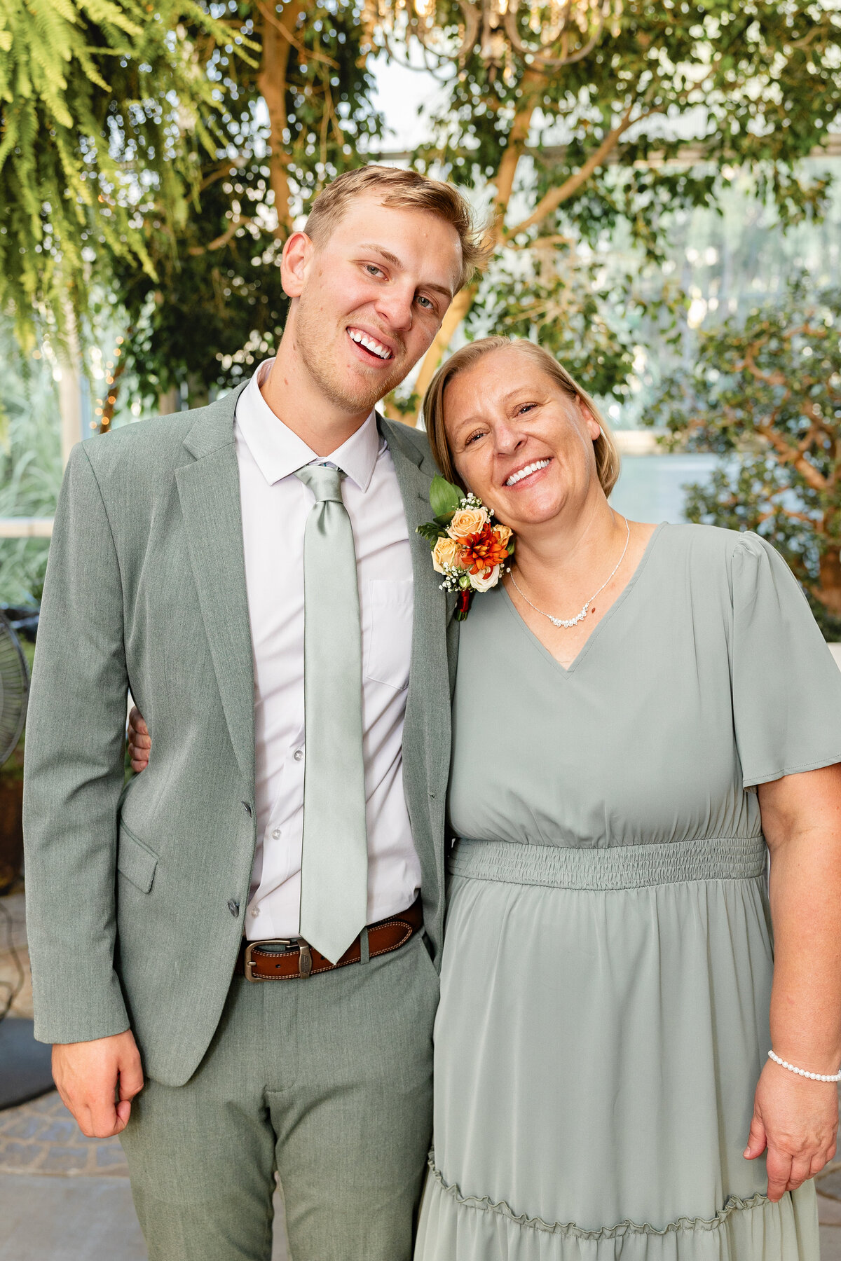 The groom, dressed in a sage green suit, smiles warmly beside his mother, who wears a matching dress, as they pose together in a lush garden venue.