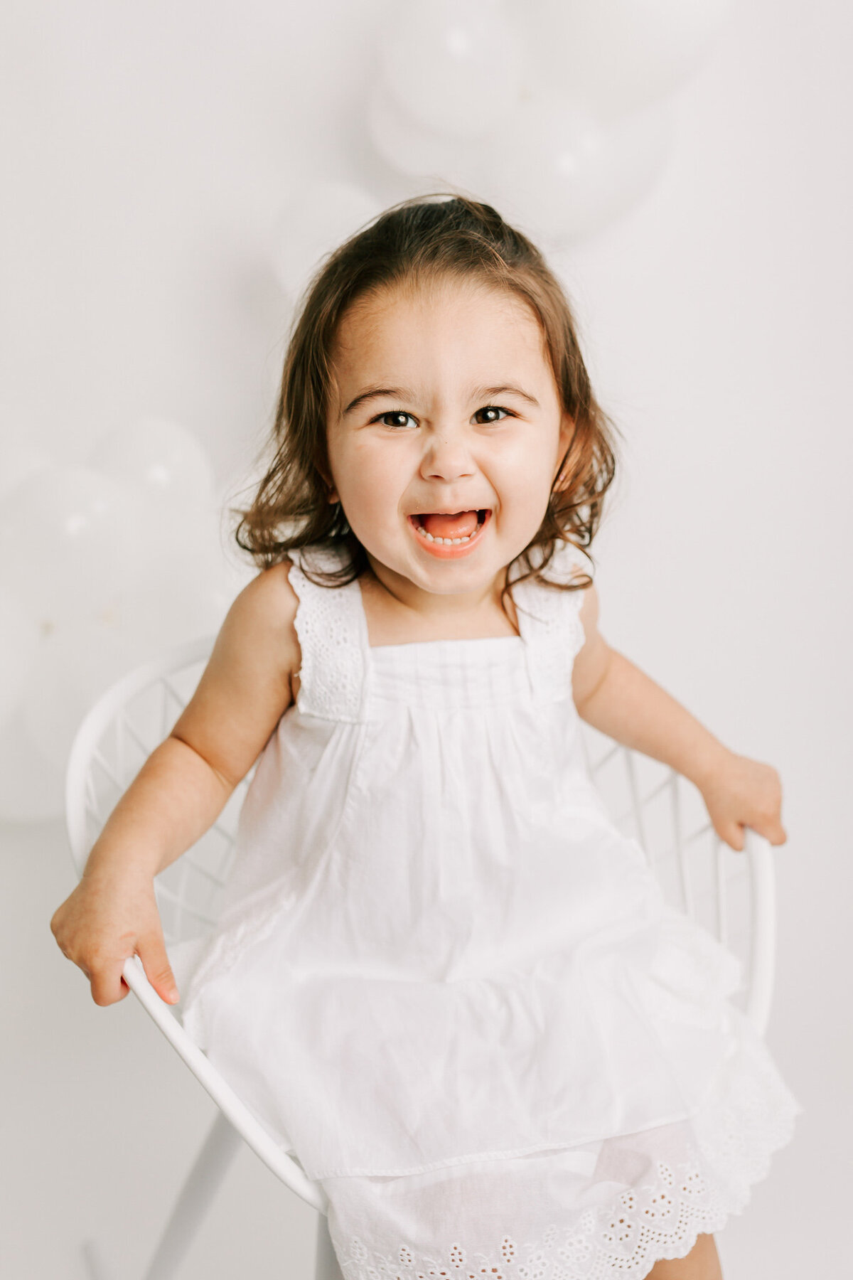 Little toddler has photos in preparation for her 2nd birthday. She sits in a white rocking chair and wears a white dress.