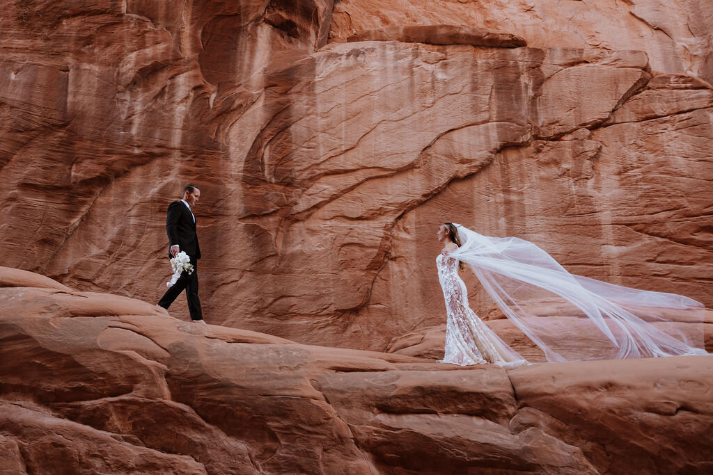 groom holding the bouquet and walking to his bride whis veil is blowing in the wind