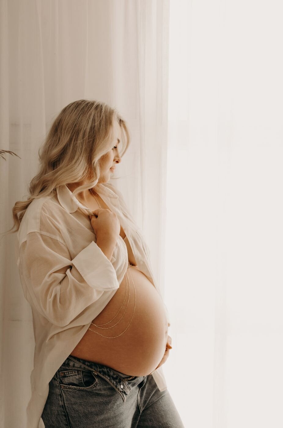 Side profile of a pregnant woman with flowing blonde waves, wearing a delicate body chain, during a professional maternity shoot by a photographer in Mullingar, Ireland.
