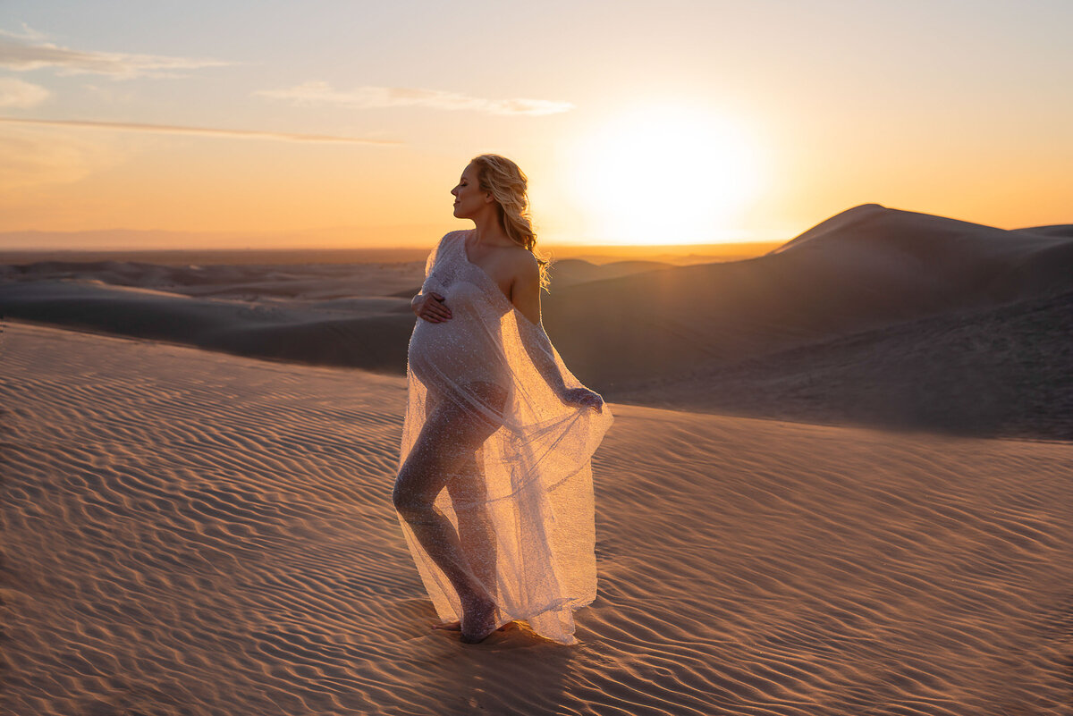 Desert maternity photoshoot by top Philadelphia maternity photographer. Expectant mom at golden hour sunset standing in the middle of the desert with the sun setting behind her. She is standing side profile to the camera with her back hand atop of her bump and forearm holding her flowing dress behind her.