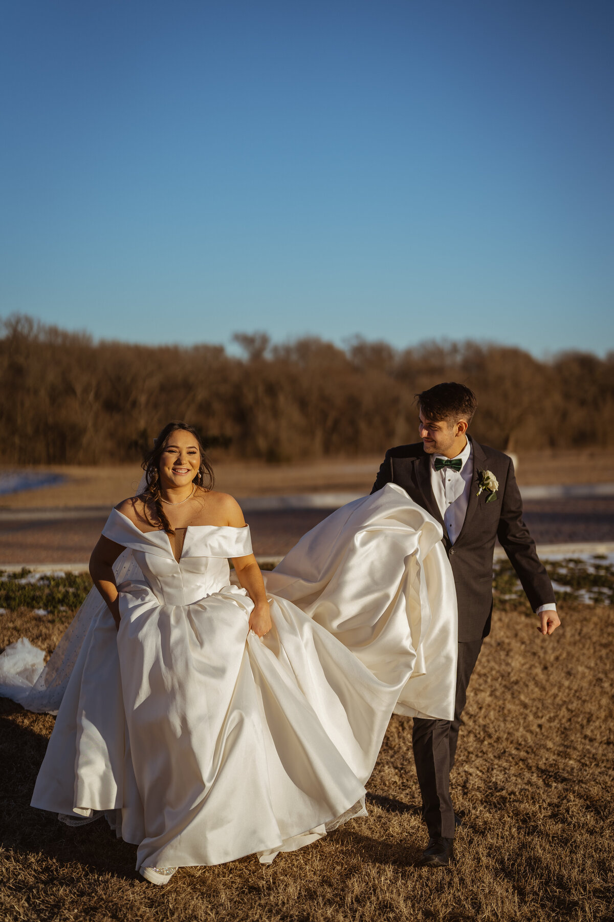 Joyful bride and groom walking together during a winter wedding at D'Vine Grace Vineyard in McKinney, Texas. The groom helps carry the bride's dress as they smile and enjoy their wedding day.