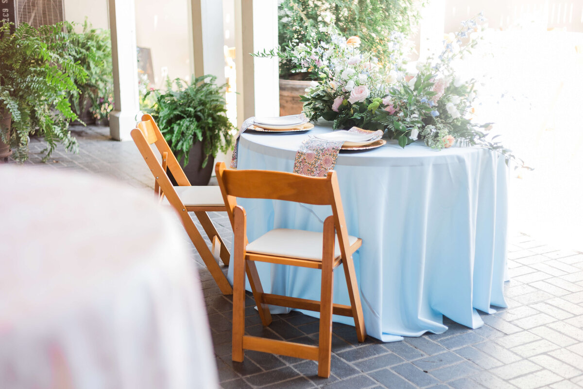 A round table covered with a light blue tablecloth is set for two with plates and napkins. A floral arrangement decorates the center. Two wooden chairs are placed at the table, and green plants are visible in the background.
