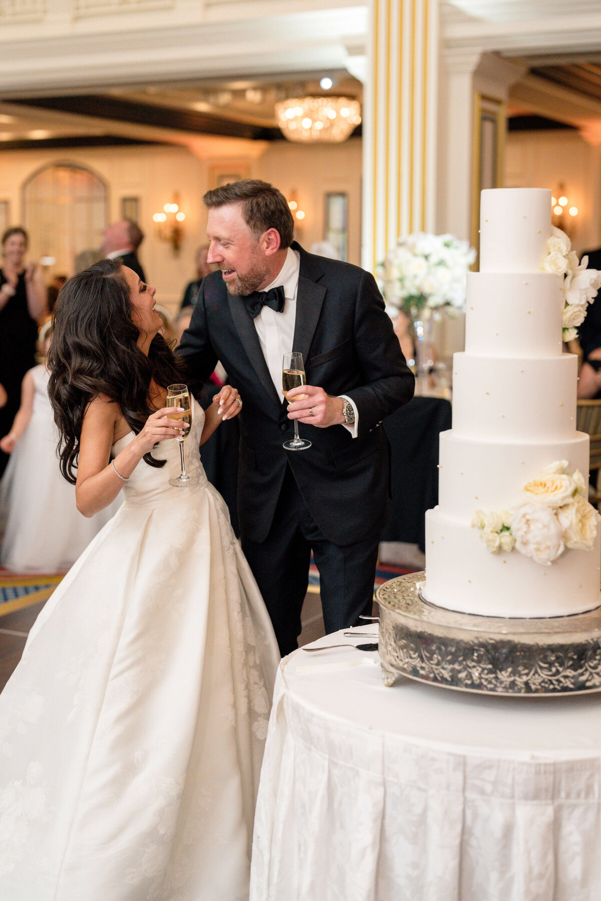 A bride in a white gown and a groom in a black tuxedo share a joyful moment while holding champagne glasses, standing near a five-tiered white wedding cake adorned with flowers. The setting is an elegant indoor space with chandeliers and guests in the background.