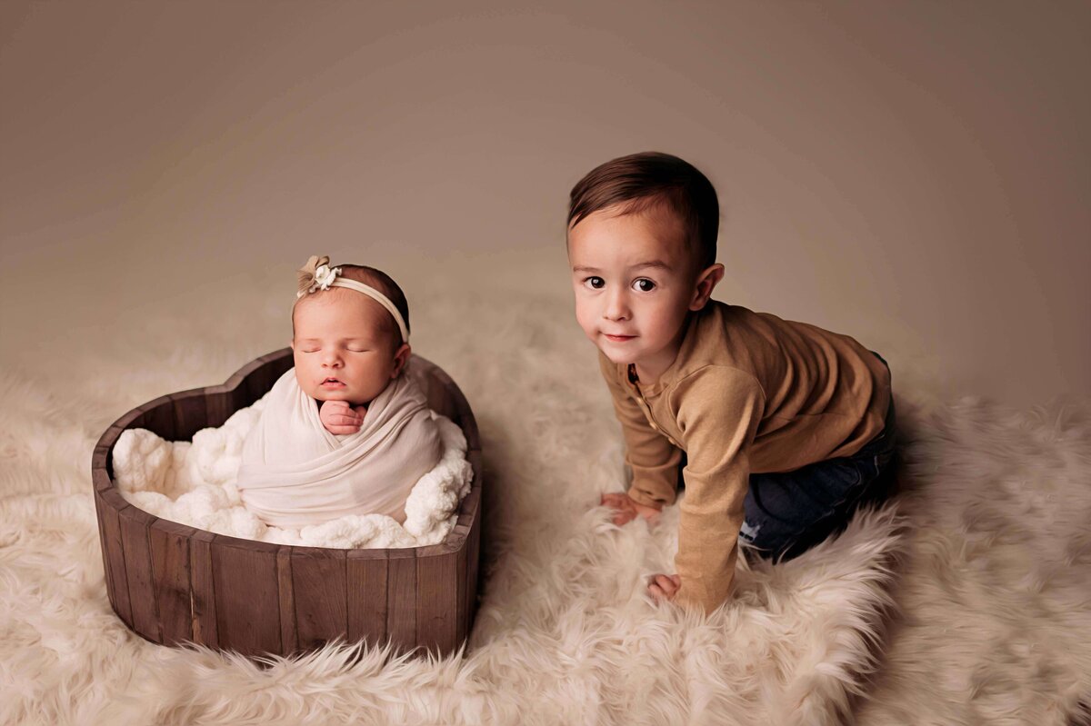 A toddler boy makes eye contact with the camera, as he sits near his sleeping newborn sister in this perfect newborn photo by Mobile, AL newborn photographer. The boy wears a brown t shirt and blue jeans, sitting a soft cream fur rug, with his newborn sister swaddled in a potato sack inside a heart bowl prop.