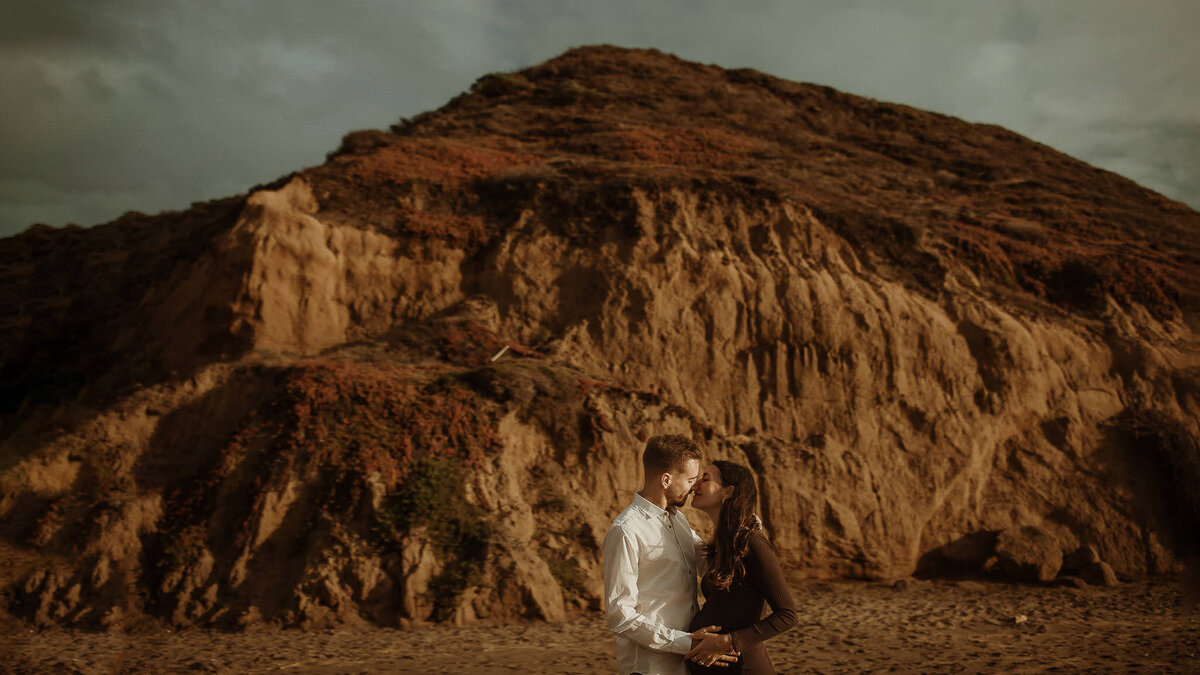 Lifestyle maternity photography session on Baker beach in SF.  Couple kisses and snuggles pregnant belly