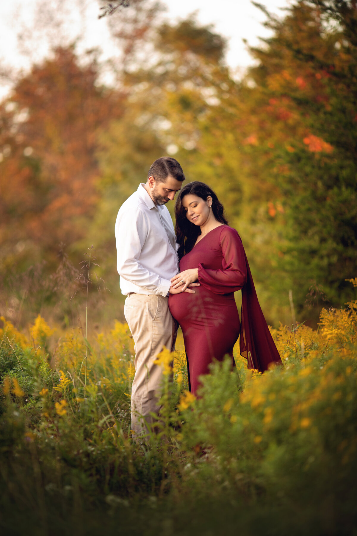 Happy expecting parents smile while resting their hands on the bump in a field of tall grasses at sunset