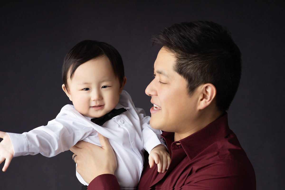 Dallas baby photographer captures a tender moment of a father and son, both smiling as they share a playful moment together.