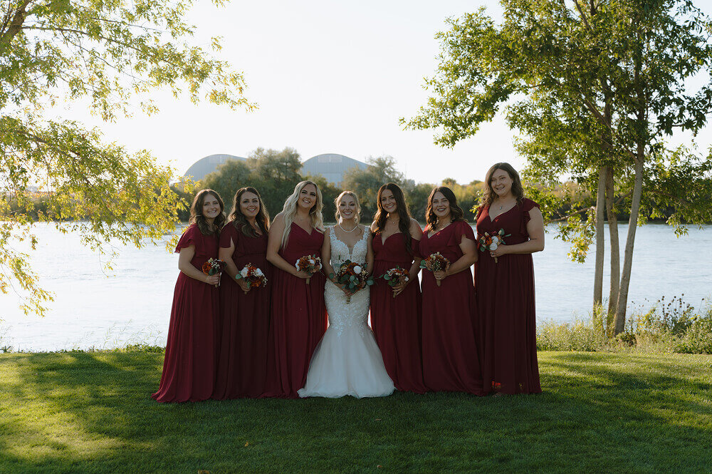 A group photo of the bridesmaids with the bride in the middle. They're wearing scarlet red dresses and standing in a grass lawn in from of a river.