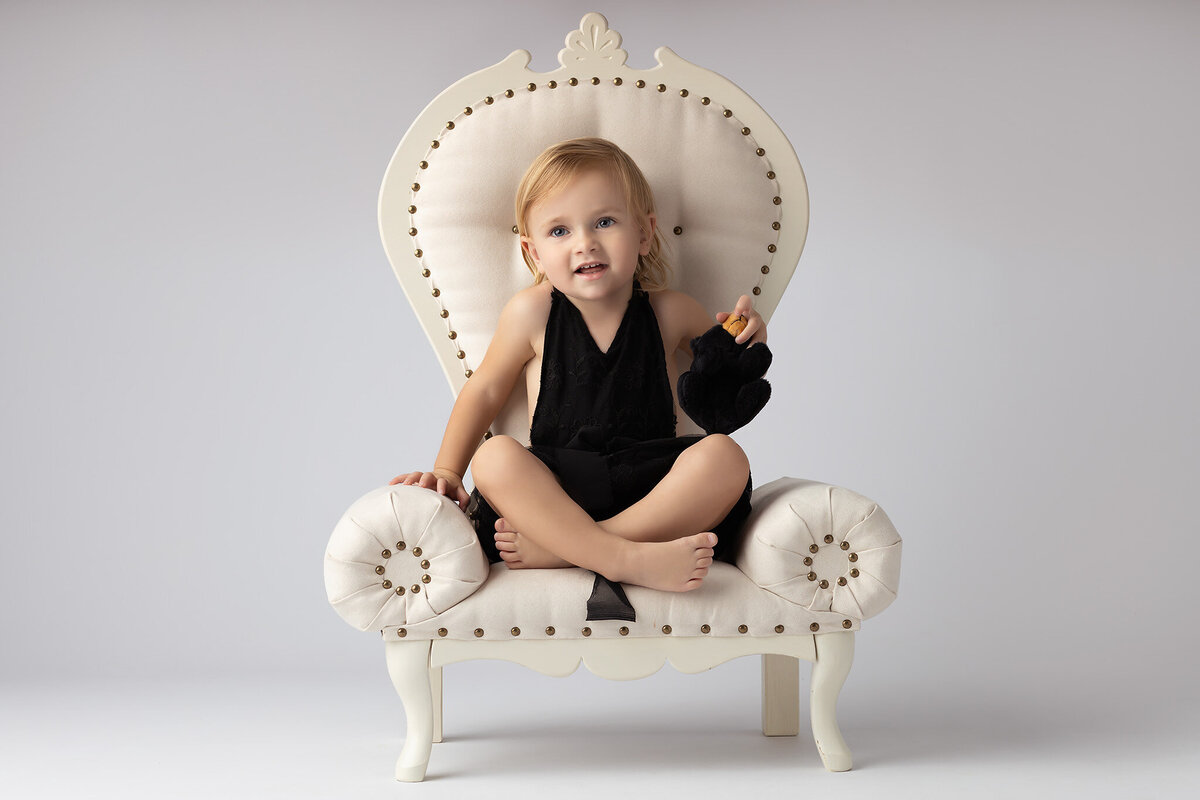 Toddler seated on a vintage chair, holding a black stuffed animal, captured by a Dallas family photographer.