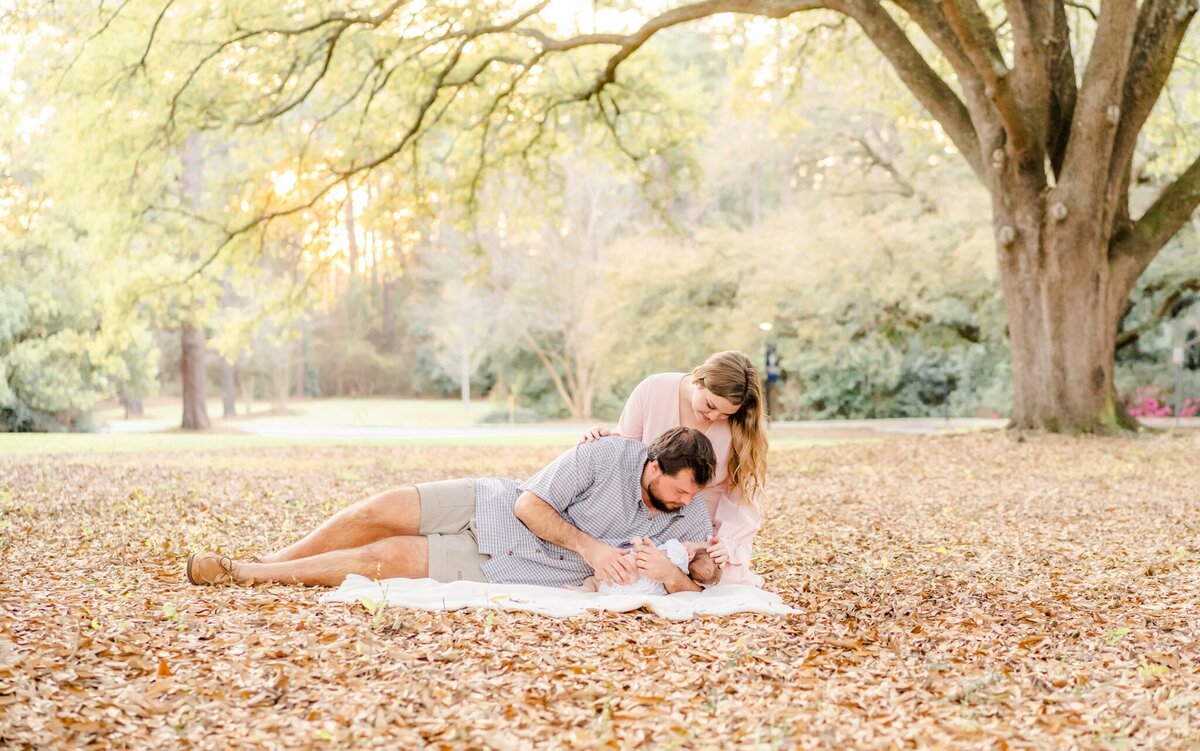 baby with parents laying on blanket admiring baby under tree at local college campus