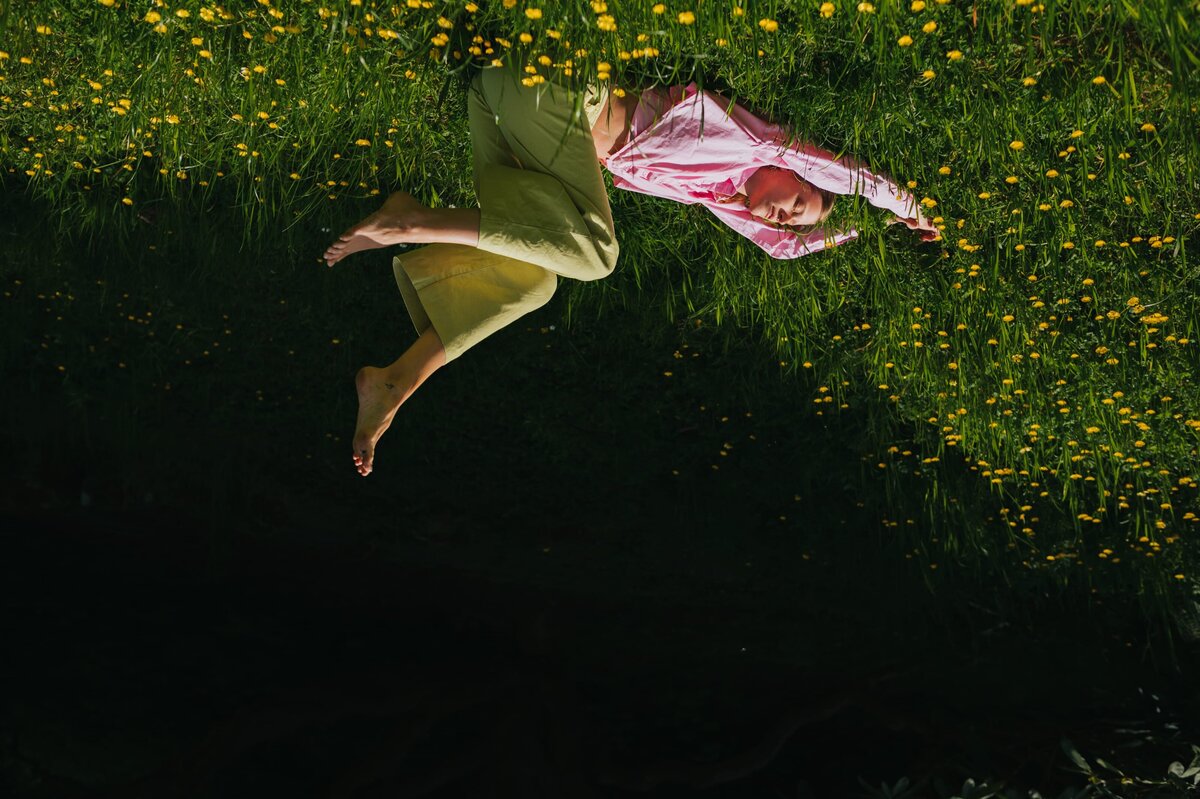 Upside down photo of a woman in a pink top and lime pants lays in a field of yellow flowers with her arms stretched overhead