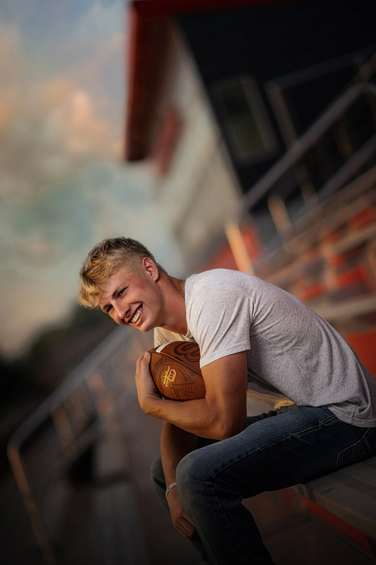 A high school senior laughs while sitting in stadium bleachers holding a football