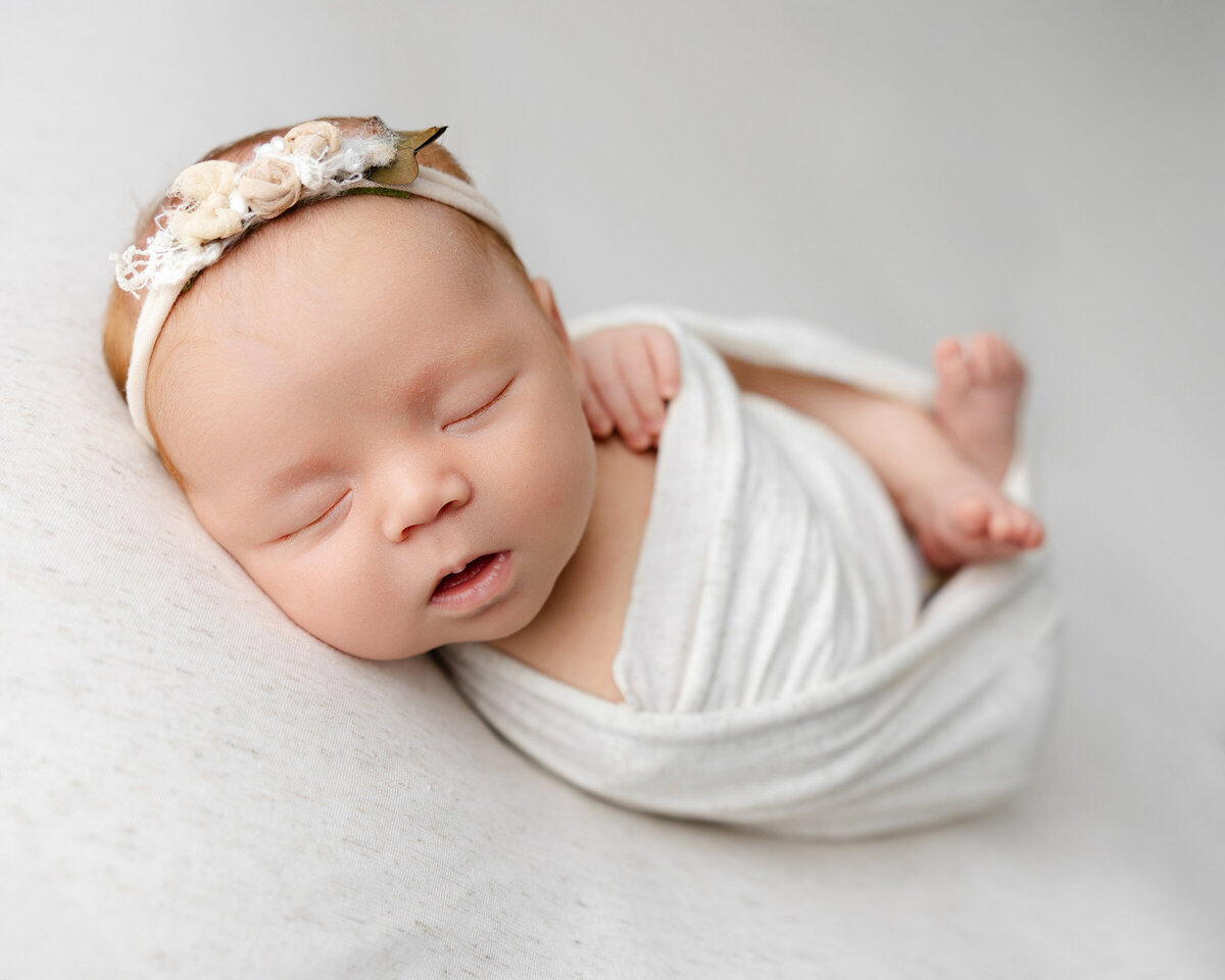 Sleeping newborn girl wrapped in a soft white swaddle with a delicate floral headband, captured during her studio session in the Inland Empire. Photograph by Kaitlyn Dawn Photography.