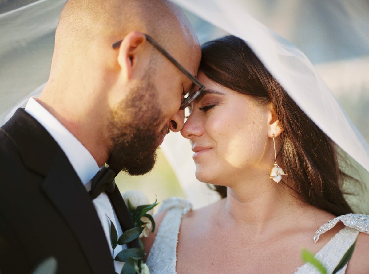 Portrait of a handsome groom in a black tuxedo touching foreheads with a dark haired bride in a white wedding dress underneath a white veil with their eyes closed.