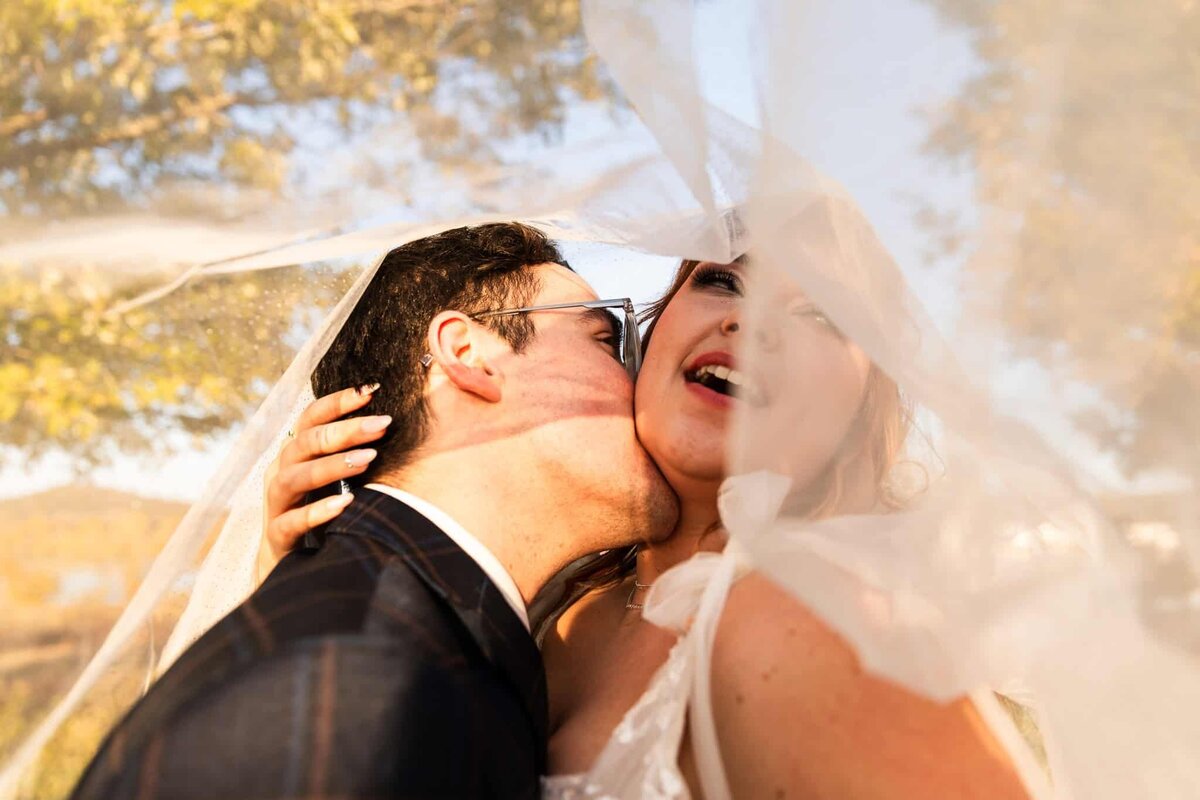 groom kisses brides neck underneath in a photo shot from beneath the wedding veil in an outdoor, autumn setting