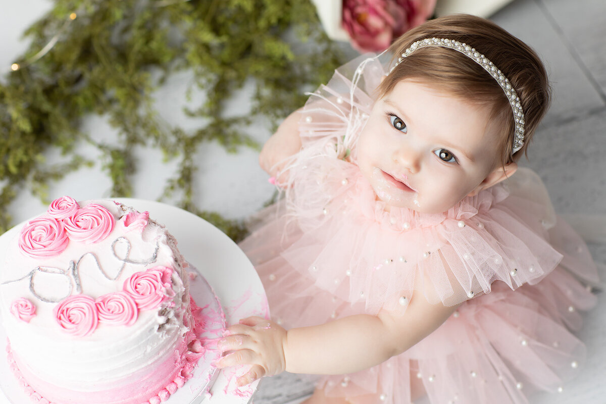 Milestone photography in Dallas featuring a baby girl in a pearl-embellished pink dress gazing up beside a pink and white cake that reads “one,” with soft greenery and roses in the background.