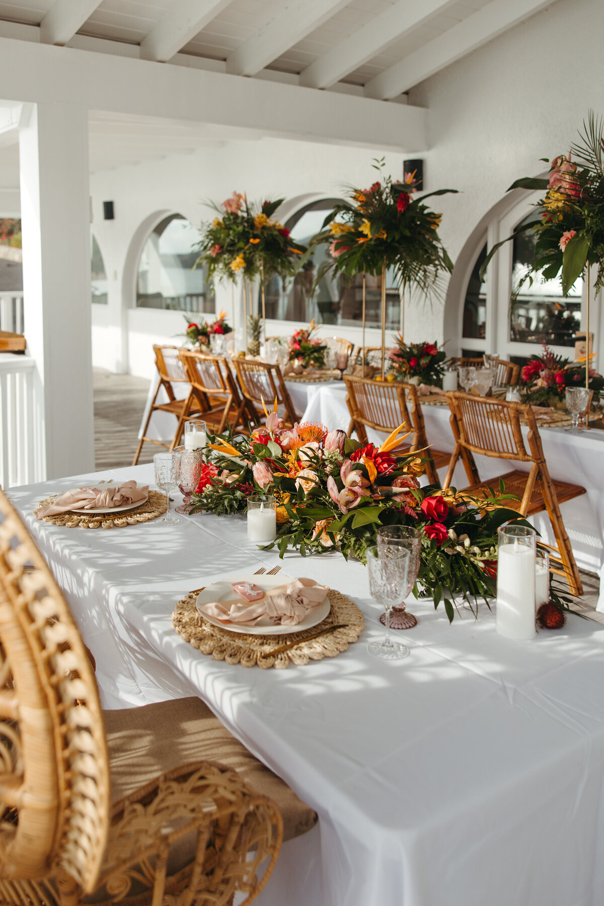 A tropical-themed wedding tablescape with vibrant floral arrangements and rattan chairs on a white linen table