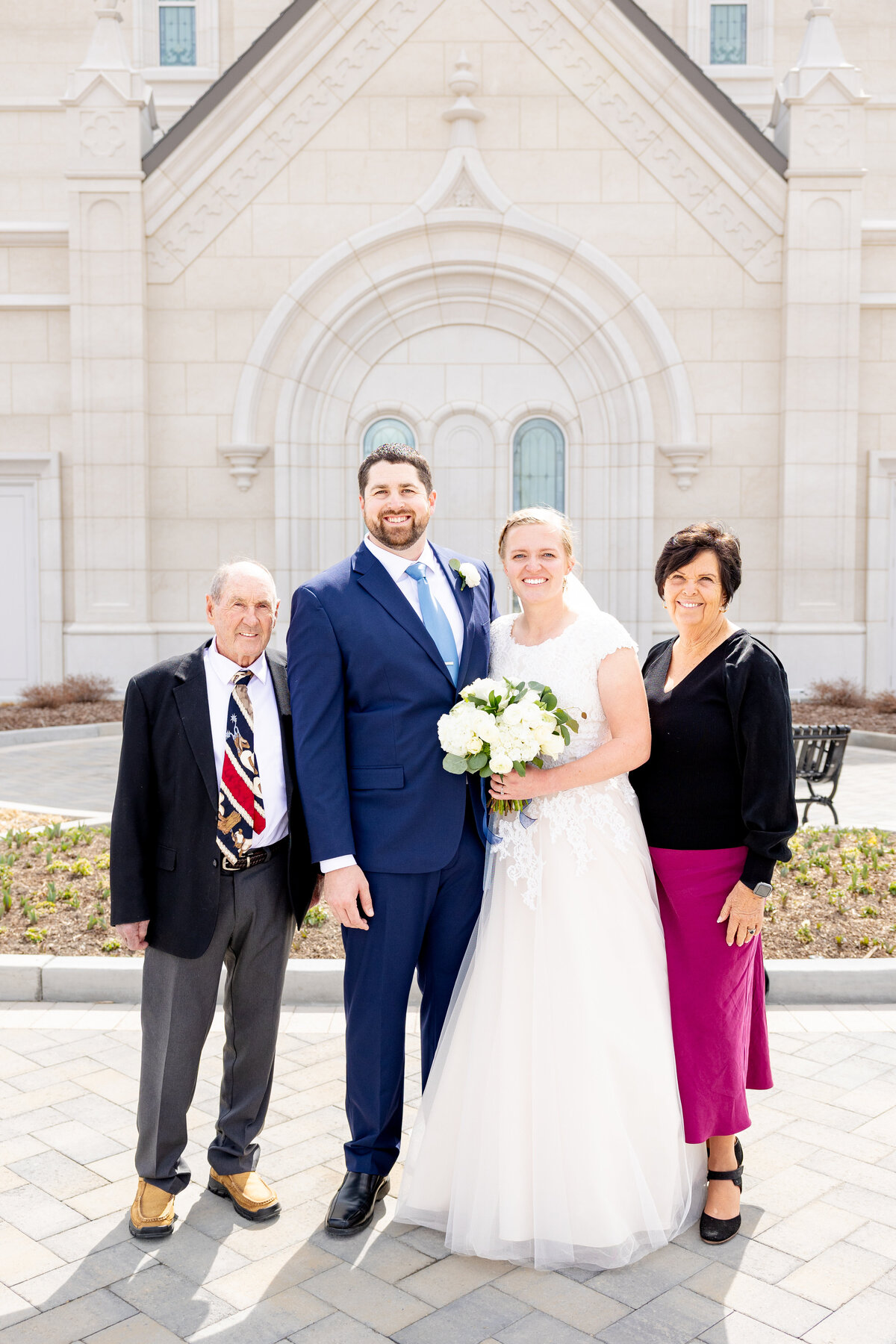 Bride and groom smile while standing arm-in-arm with the groom’s grandparents, in front of the Taylorsville Utah LDS Temple on a sunny day.
