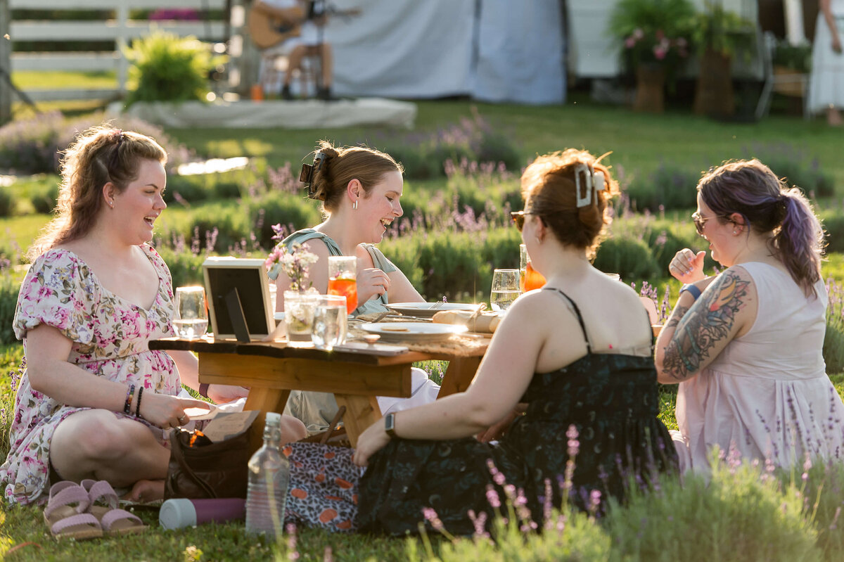 women laughing and sitting at a picnic charcuterie as part of Soiree in the Field.  Captured by Ottawa Event Photographer JEMMAN Photography COMMERCIAL