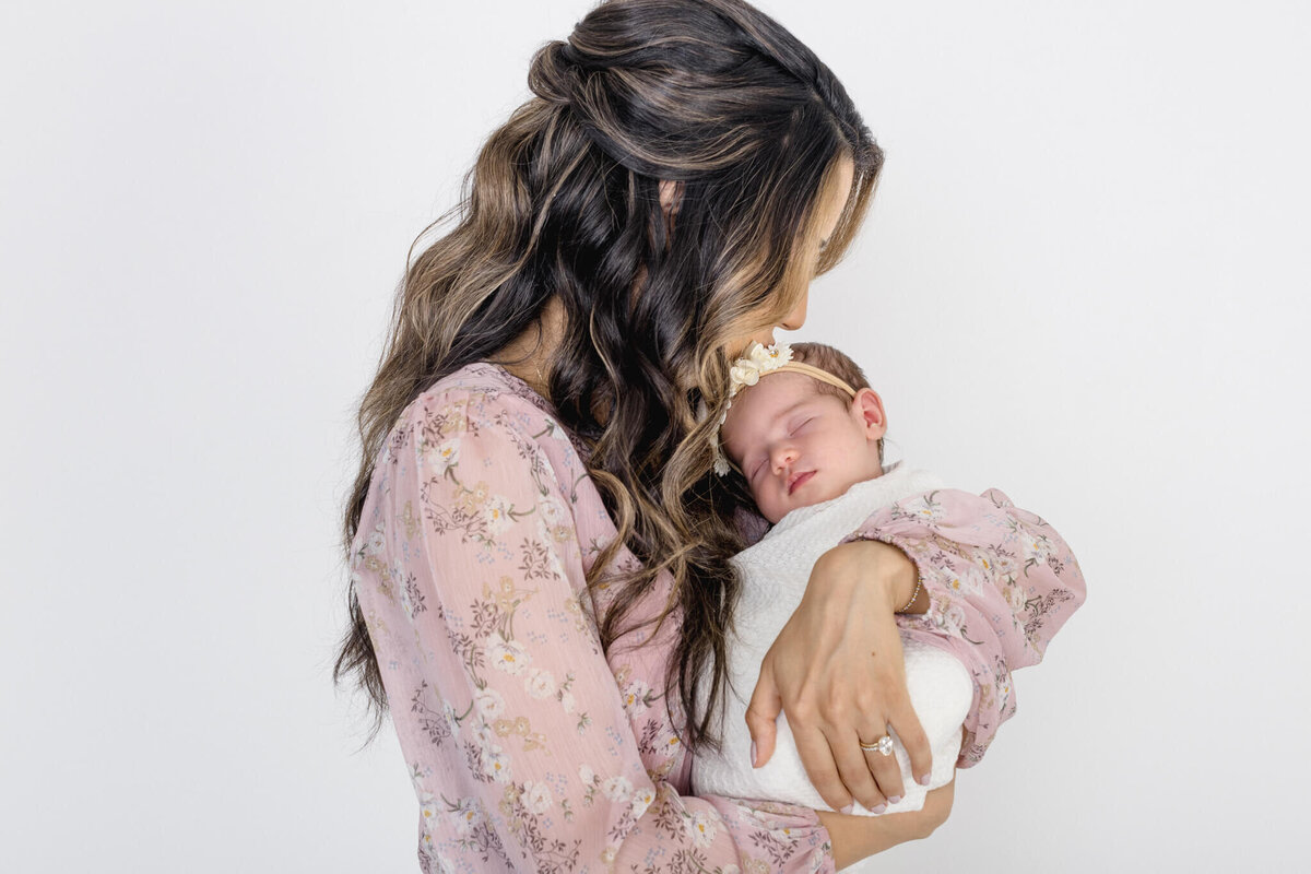 A woman with long, wavy hair in a floral dress gently cradles a sleeping baby swaddled in white. The baby wears a headband with a bow. They both exude a serene and tender moment against a plain white background.