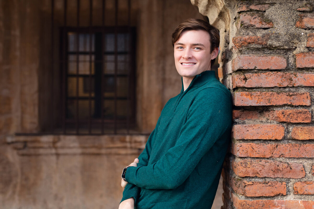 A young man with brown hair wearing dark green smiling at the camera and leaning against a brick wall.