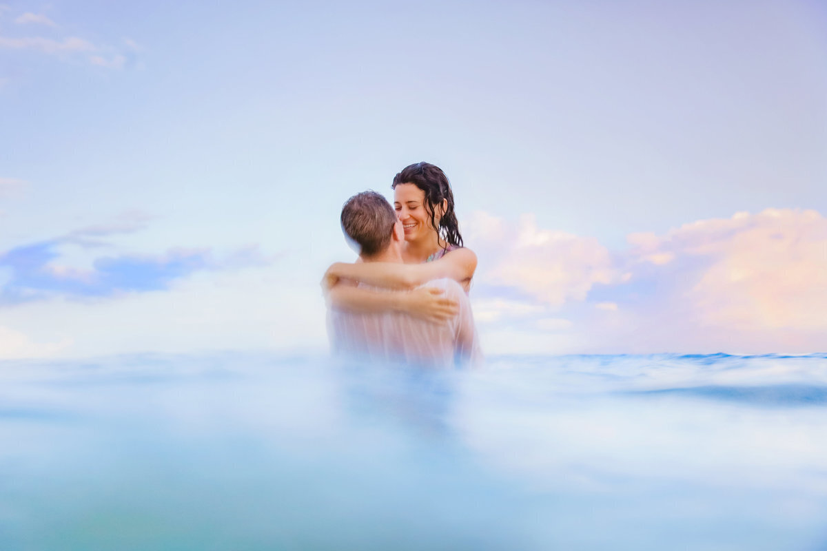 Brunette woman wraps her arms around her husband while the two stand halfway in the water off Wailea Beach in Maui