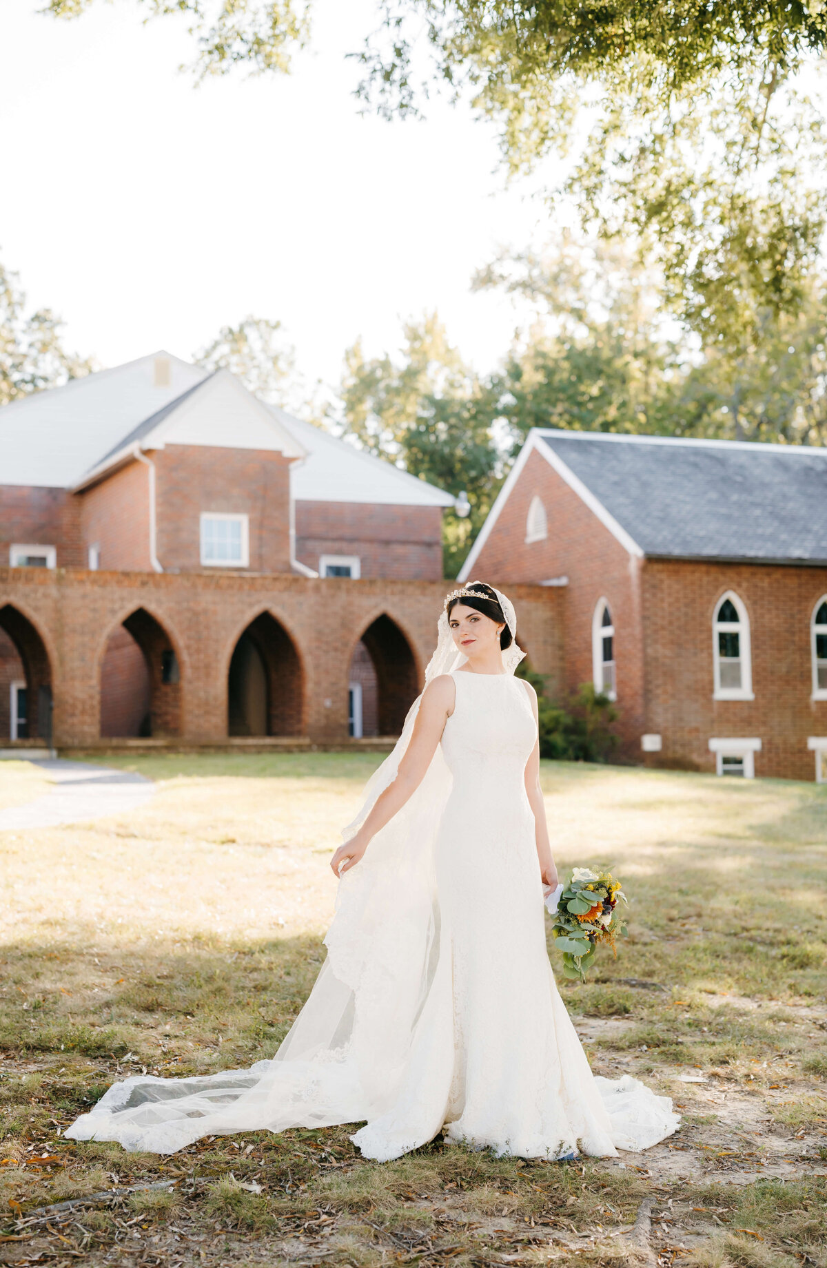 brick wedding chapel stands behind bride in a 1920s style wedding dress with modern touches as she poses with her hand gently holding her cathedral length wedding veil to the side and her wedding bouquet with her other hand for elegant wedding photos