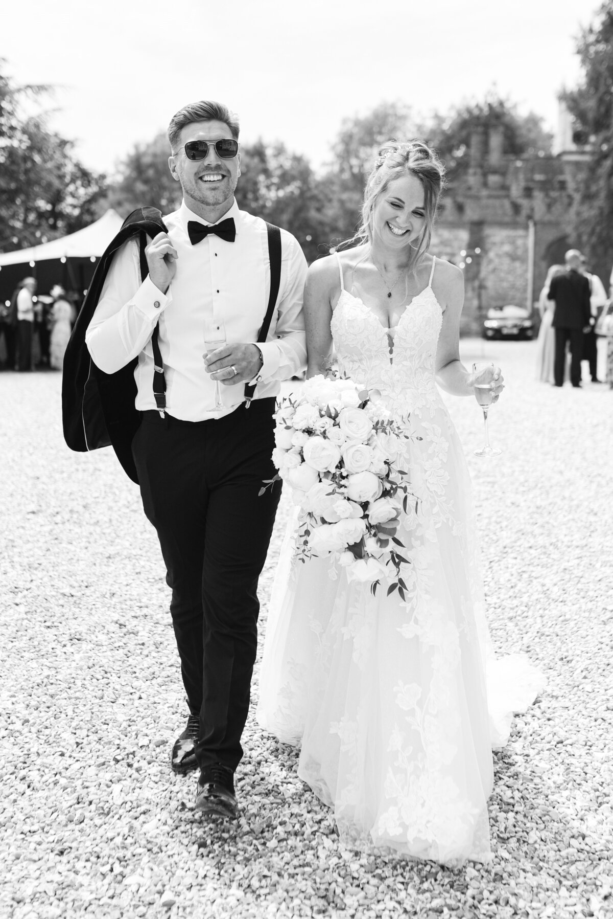 Black and white image of a bride and groom, they are walking together smiling. the groom is holding his jacket over his shoulder and is wearing sunglasses, the bride is wearing a white lace dress with thin straps and is holding her bouquet and a glass of champagne