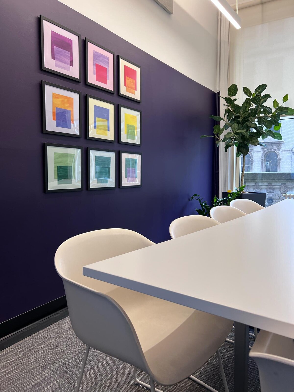Bright meeting space with purple accent wall and a grid of nine colorful abstract framed prints.