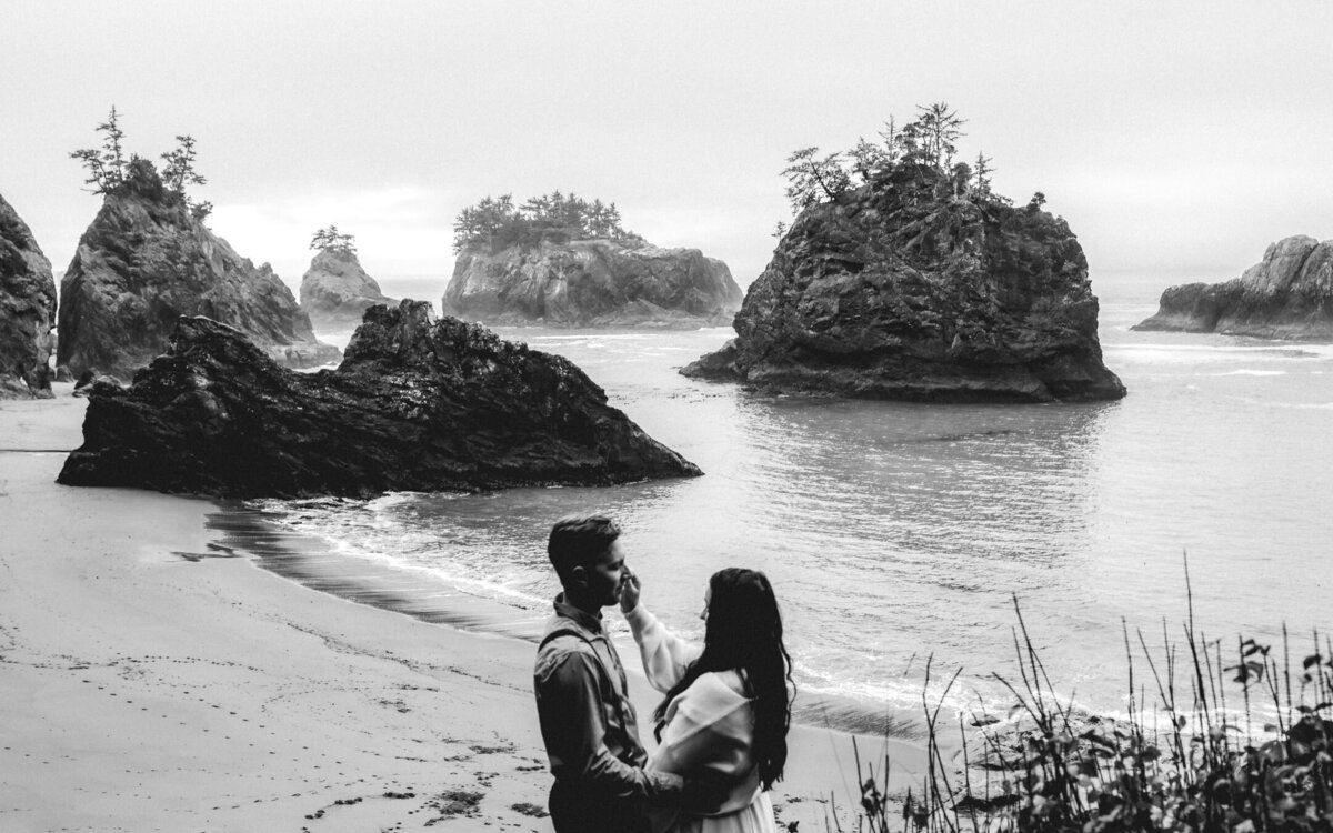 during their oregon coast elopement, a couple stands above a moody beach. She strokes his cheek as  he gazes lovingly at her.