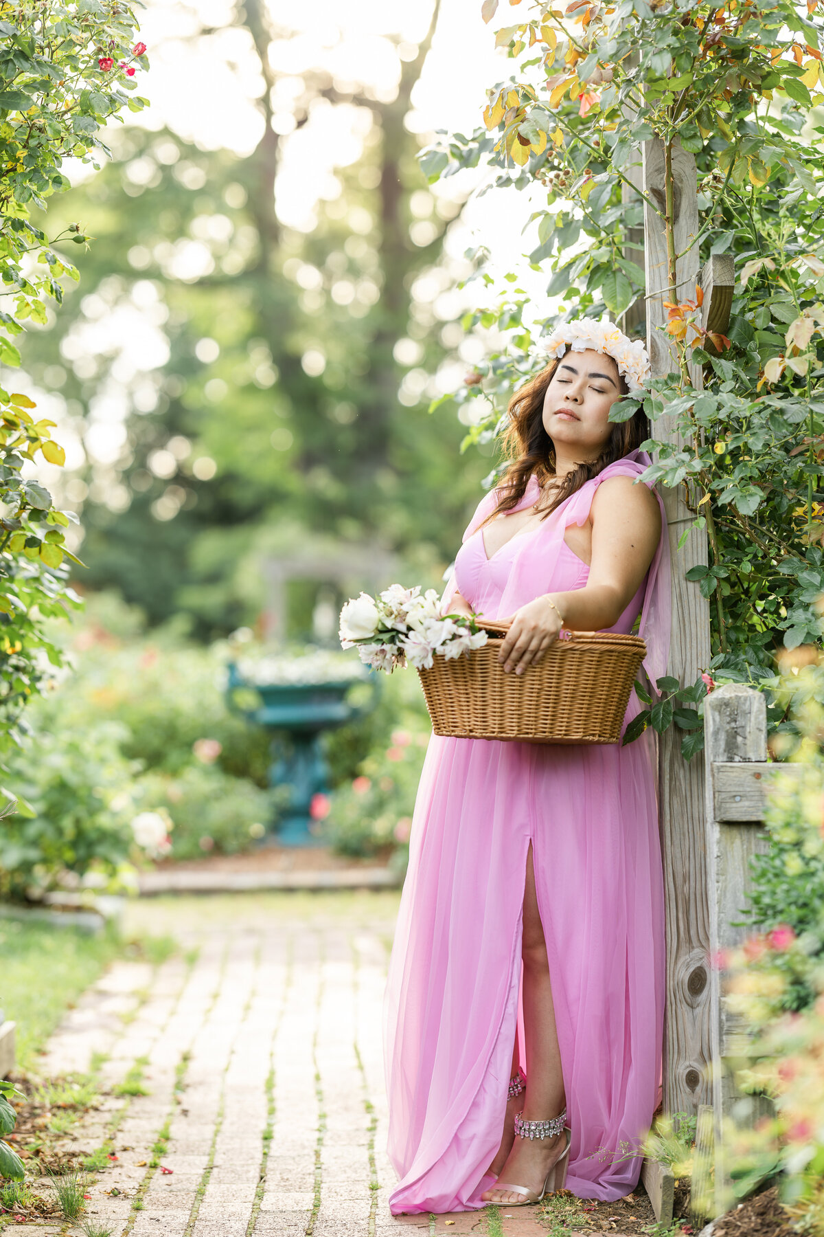 A young lady wearing a pink gown, leans against the archway of a vibrant garden while holding a basket of flowers. Her eyes are closed with her head tilted upward as she soaks in the beauty of the garden that surrounds her. 