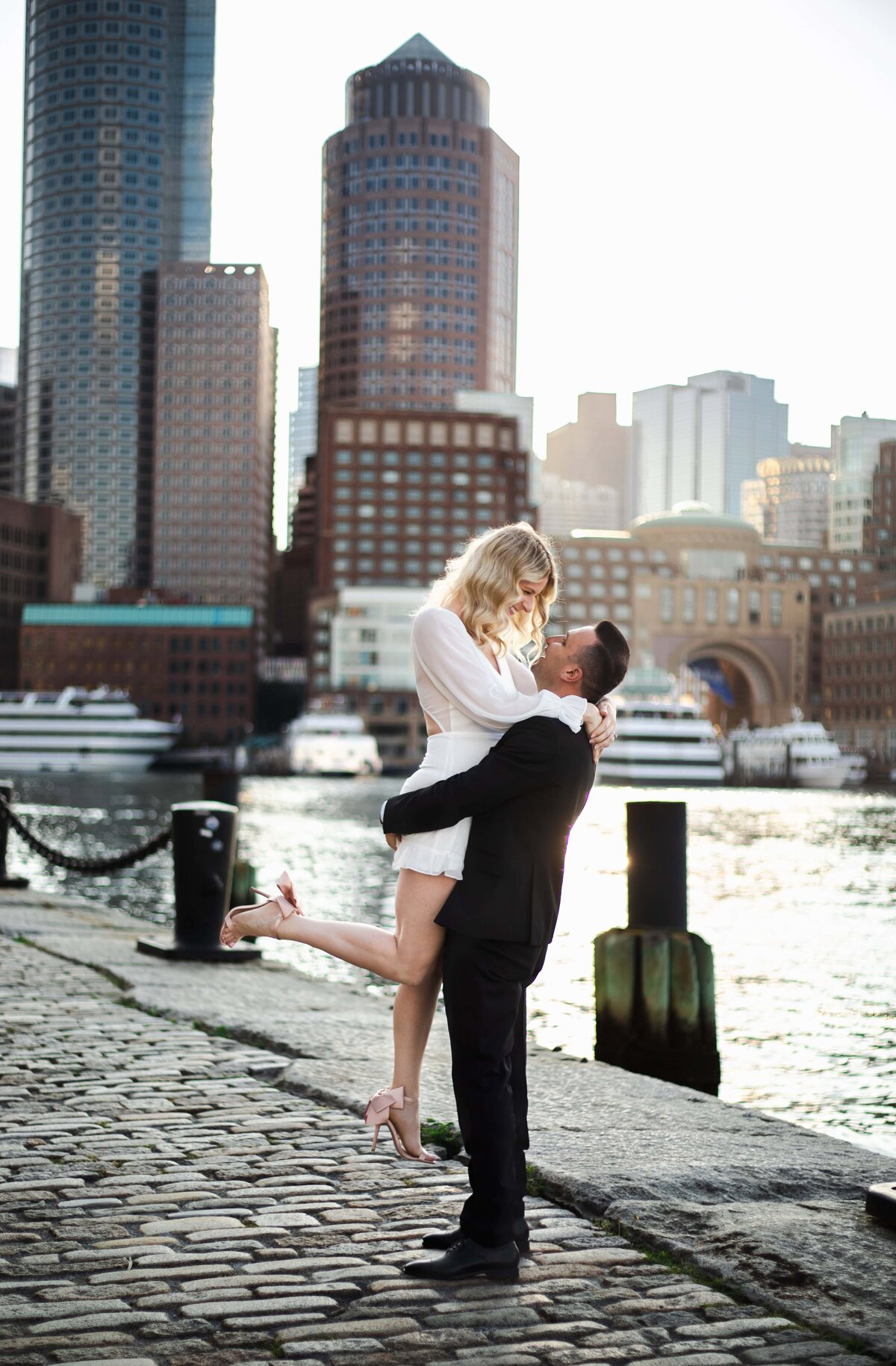 Engagement Photos Rhode Island | Kelsey Sheehan Photography Timeless Rhode Island Weddings | A couple embraces by the waterfront, the woman in white lifted by a man in a black suit. City skyline and boats in the background, conveying romance.