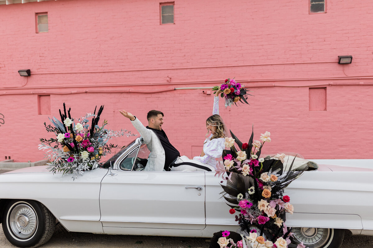 bride and groom scream with joy in their convertable car covered in floral arrangements