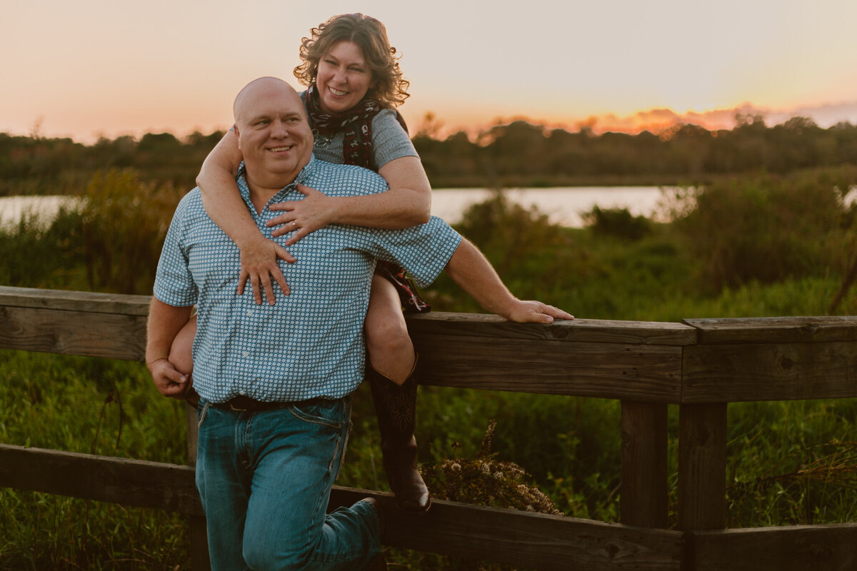 Playful engagement photo of Chad and Shane at Bay Area Park, Houston, TX. The couple is sitting on a wooden fence at sunset, laughing and embracing. Captured by ShutterUp Photography.