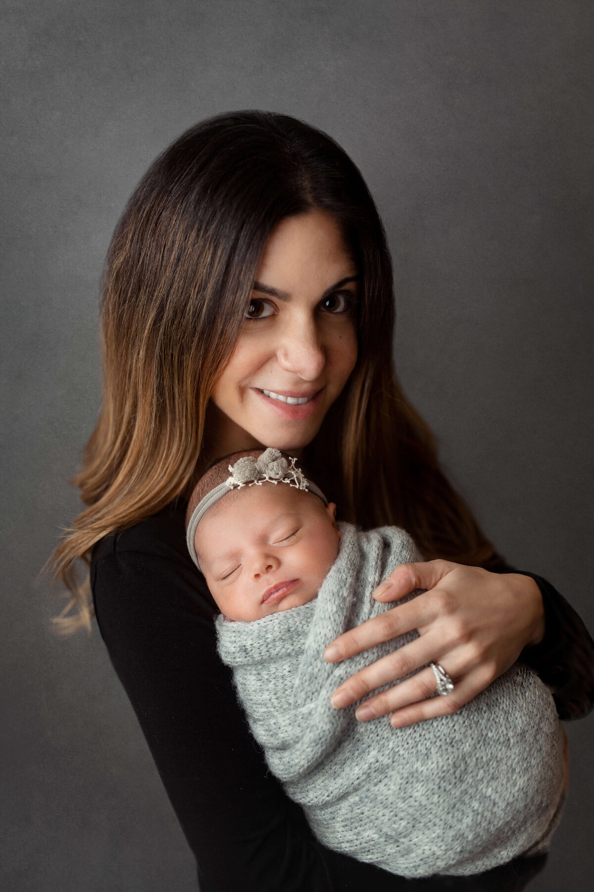 new mother wearing black holding her newborn baby girl who is swaddled in a grey knit wrap with a pretty headband during their newborn session