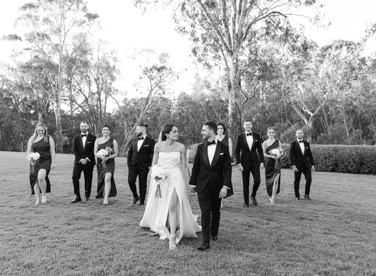 A black and white image of a wedding party in a grassy field. The bride and groom are  the focus of the image.  The groomsmen and bridesmaids follow slightly behind them.