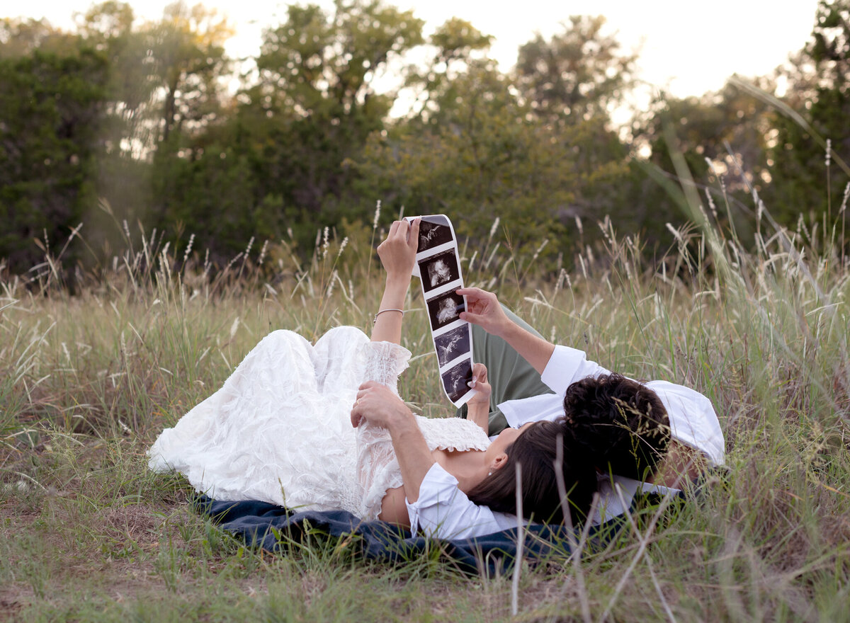 Couple lying in tall grass holding ultrasound photos during outdoor maternity session in Georgetown, TX