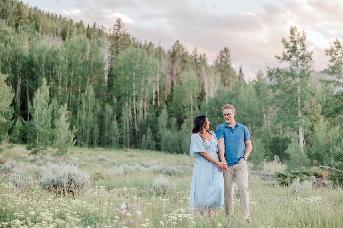 A happy couple in blue giggles while walking in a mountain meadow holding hands