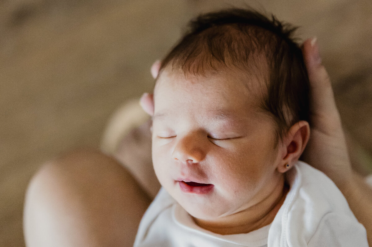 A newborn baby with dark hair sleeps peacefully while being cradled in someone's hands.